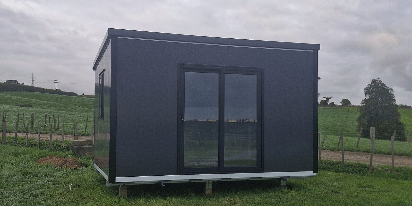 A 4 × 3 m charcoal Simple Cabins cabin with a ranch slider, on piles in a paddock