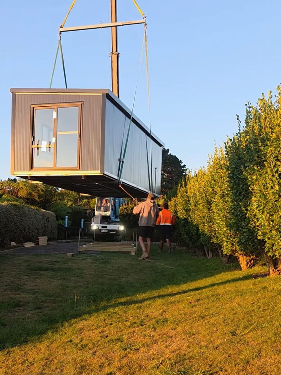 A cabin craned over a hedge into a back garden in late afternoon light