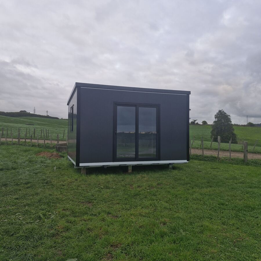 A 4 × 3 m charcoal cabin with a ranch slider, sited on piles in a rural paddock